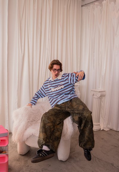 Joven con camisa de rayas y pantalones de camuflaje sentado en una silla blanca esponjosa en una habitación con cortinas blancas y columnas clásicas.