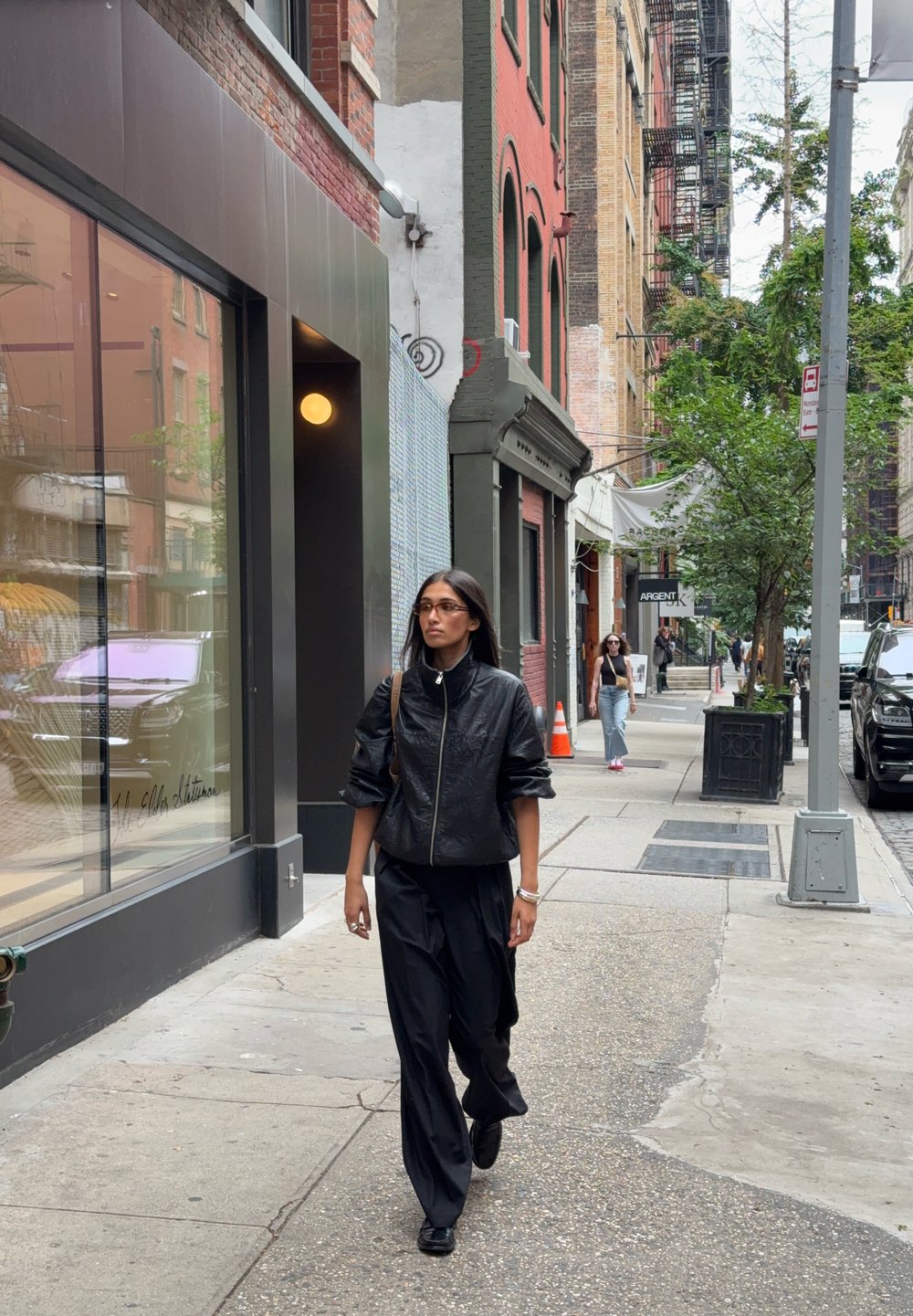 Chaqueta bomber negra con un cuello alto, tela texturizada, combinada con pantalones negros sueltos y zapatos negros, caminando por la acera de una ciudad.