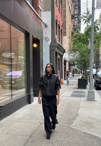 Chaqueta bomber negra con un cuello alto, tela texturizada, combinada con pantalones negros sueltos y zapatos negros, caminando por la acera de una ciudad.