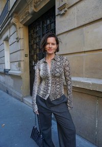 Långärmad skjorta med ormtryck i beige och brunt, med en figurnära design och knappstängning. Matchad med lösa grå byxor och en svart handväska.