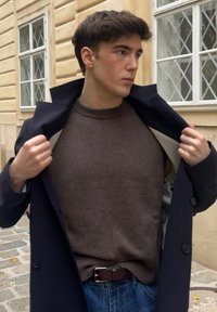 Marineblå lang frakk over en brun strikkegenser med rund hals. Lyse denimjeans og et brunt belte, utendørs nær en teksturert vegg.