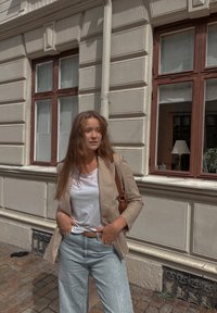 Ανοιχτό μπεζ σακάκι πάνω από λευκό μπλουζάκι, σε συνδυασμό με ανοιχτό τζιν. Καφέ τσάντα ώμου, μπροστά από ένα κτίριο με παράθυρα.