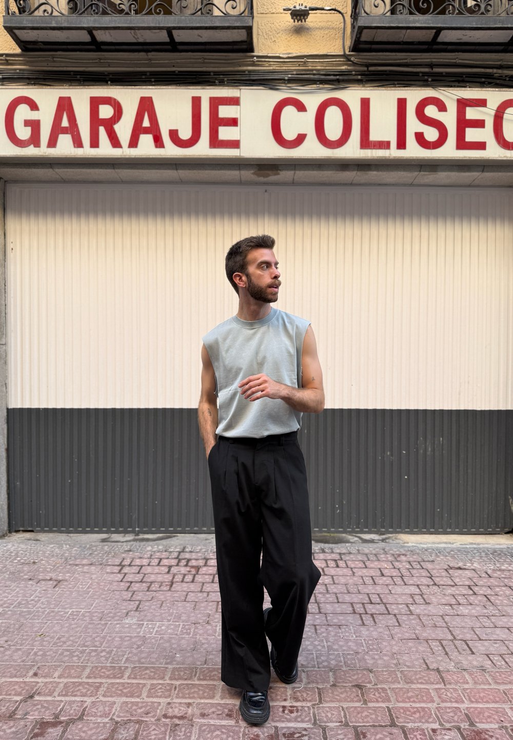 Gaiši pelēks bez piedurknēm tops, apvienots ar brīvām melnām biksēm un melnu apavu; stāv uz teksturētas ietves ar neitrālu fonu.