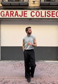 Gaiši pelēks bez piedurknēm tops, apvienots ar brīvām melnām biksēm un melnu apavu; stāv uz teksturētas ietves ar neitrālu fonu.