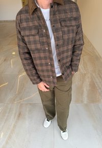 Brun ternet jakke med en ribbetrøje-krave, grå t-shirt, olivengrønne cargobukser og hvide sneakers, vist på et marmorgulv.