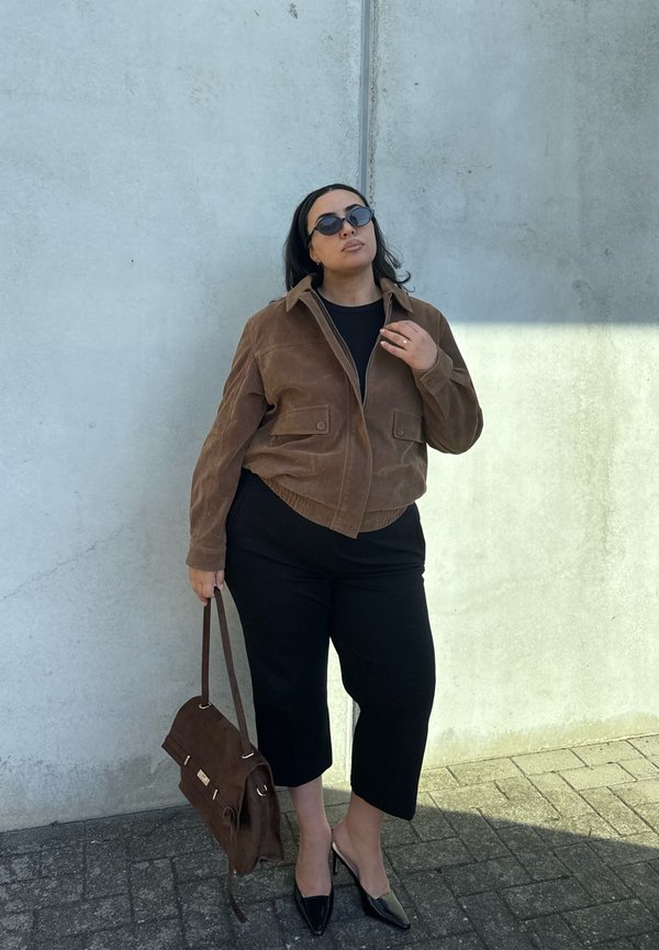 Una mujer con gafas de sol, una chaqueta marrón, pantalones negros y zapatos de tacón posa junto a un muro de hormigón, sosteniendo un bolso marrón de hombro.