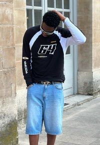 Camisa preta de manga longa com mangas brancas e detalhes gráficos em dourado, combinada com calções de ganga azul-claro e um cinto com um logo metálico.