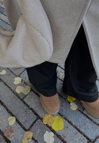 Beiger, flauschiger Beutel, der über einen hellgrauen Mantel gelegt ist; dunkle Hose und beigefarbene Wildlederstiefel auf einem Kopfsteinpflasterweg mit verstreuten gelben Blättern.