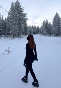 Personne en tenue d'hiver noire et bottes marchant sur une route forestière enneigée entourée de grands pins couverts de neige sous un ciel nuageux.
