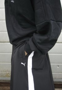 Schwarzer Pullover mit Rundhalsausschnitt und schwarze Hose mit weißen Seitenstreifen und Puma-Logo, mit den Händen in den Taschen.