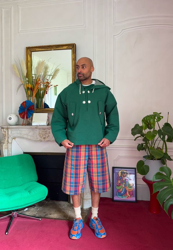 Bărbat purtând o jachetă verde, pantaloni scurți în carouri și adidași colorați stă într-o cameră cu un scaun verde, o plantă în ghiveci și o lucrare de artă încadrată pe podea.