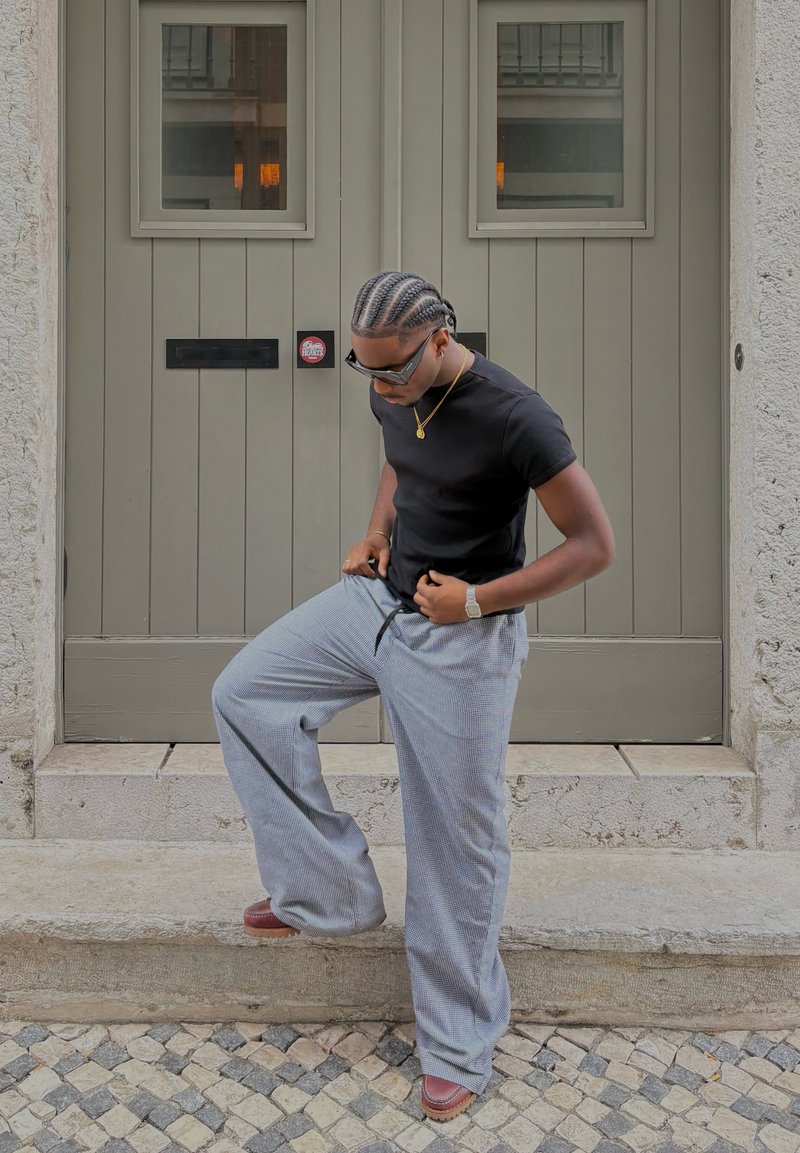 Must have lühivarrukaline must särk, hallid ruudulised laiade jalgadega püksid, prillid ja õhuke kuldne kett, seismas kivist treppidel, taustal uks.