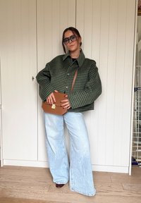 Zelená károvaná bunda, svetlomodré džínsy s širokými nohavicami a hnedá kabelka na rameno. Textúrovaná látka, voľný oversized strih a dlhé rukávy so zapínaním na gombíky.