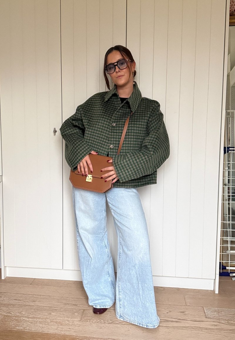 Casaco verde com padrão xadrez, jeans azul claro de perna larga e mala castanha de ombro. Tecido texturizado, corte oversized e mangas compridas com fecho de botões.