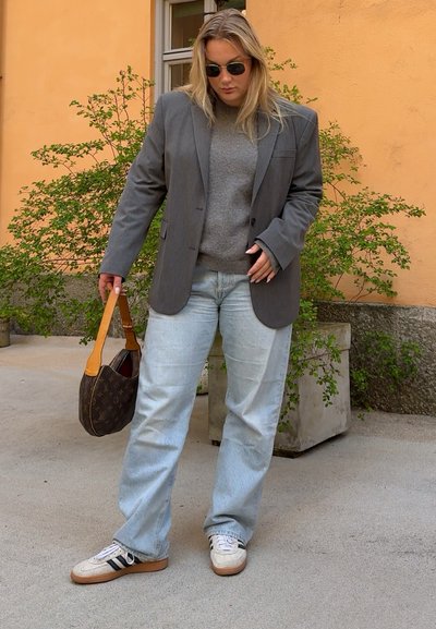 Blazer gris oversize sobre un suéter gris, jeans baggy de color azul claro y zapatillas blancas con rayas negras. Bolso marrón con detalles en color marrón claro.