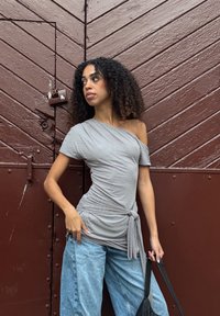 Sivi top s otvorenim ramenima i suptilnim točkastim uzorkom, vezan sa jedne strane, uparen sa svijetloplavim trapericama širokih nogavica i crnom torbom na rame.