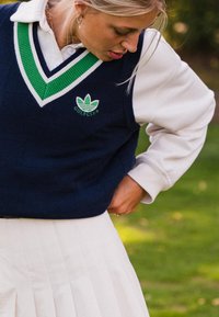 Blondīne sieviete, kas pielāgo tumši zilu golfa vestu ar zaļām svītrām, valkājot baltu kreklu un plīstošu balto svārku uz zāles fona.