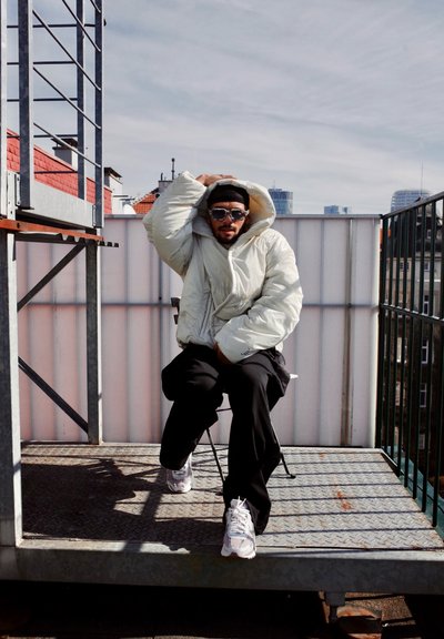 Chaqueta puffy de color claro con una capucha grande, llevada sobre pantalones negros. Persona sentada en una plataforma de metal, usando gafas de sol y zapatillas.