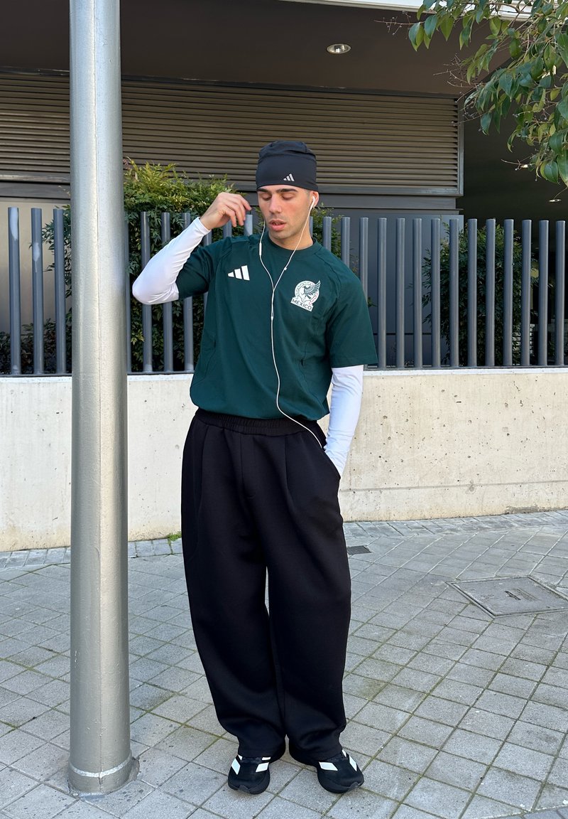 Jeune homme portant un t-shirt vert foncé du Mexique, un bonnet noir et un pantalon noir, ajustant ses écouteurs en extérieur sur une zone pavée près d'un poteau métallique.
