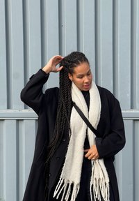 Crni velikih dimenzija kaput s velikim gumbima, bijeli teksturirani šal prebačen oko vrata i pletena kosa, na sivom ozadju.