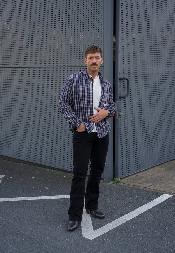 Hombre con bigote que lleva una camisa de cuadros, camiseta blanca, pantalones negros y botas, está de pie en la calle junto a una pared de metal perforado y una puerta en la esquina.