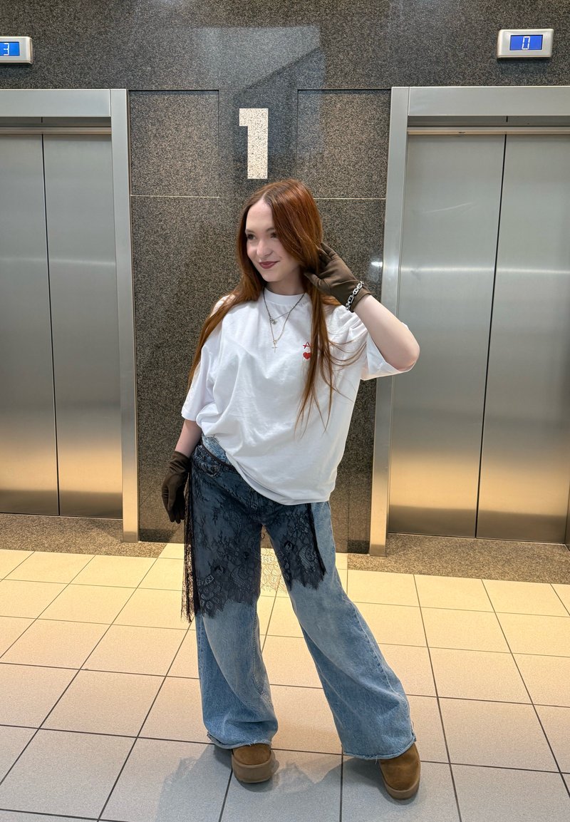 Jeune femme aux longs cheveux roux, portant un t-shirt blanc oversize, un jean ample avec de la dentelle noire, des gants marron et des bottes, se tient entre les portes fermées de l'ascenseur.