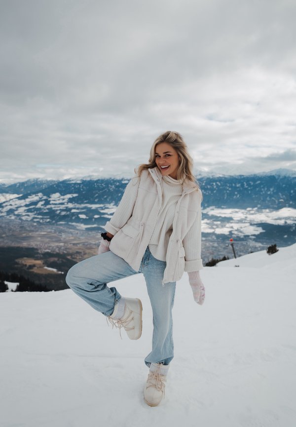Νεαρή γυναίκα με λευκό χειμερινό μπουφάν, τζιν και μπότες, που χαμογελά και σηκώνει ένα πόδι σε χιονισμένο βουνό με συννεφιασμένο ουρανό και μακρινή κοιλάδα.