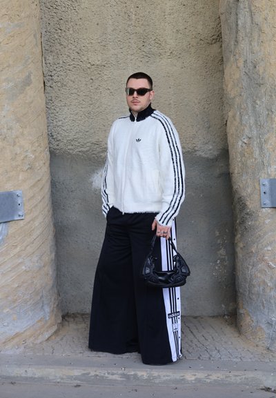 Hombre con chaqueta blanca de Adidas y pantalones anchos negros de Adidas sosteniendo un bolso negro, de pie entre pilares de piedra con textura.