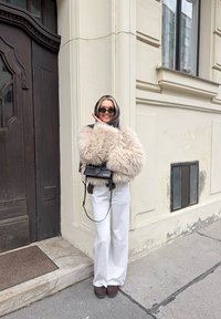 Løs, kremfarget fuskepelsjakke med vide ermer, hvite bukser og svart håndveske. Mørke solbriller og et svart hodeplagg er også godt synlige.