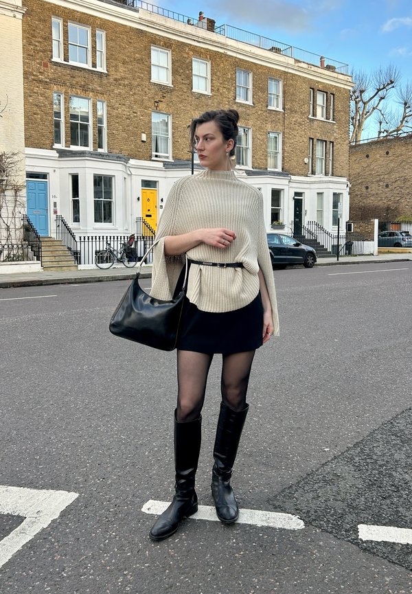 Donna in piedi per strada che indossa un poncho beige lavorato a maglia, minigonna nera, collant, stivali al ginocchio, e tiene una borsa nera con edifici in mattoni sullo sfondo.