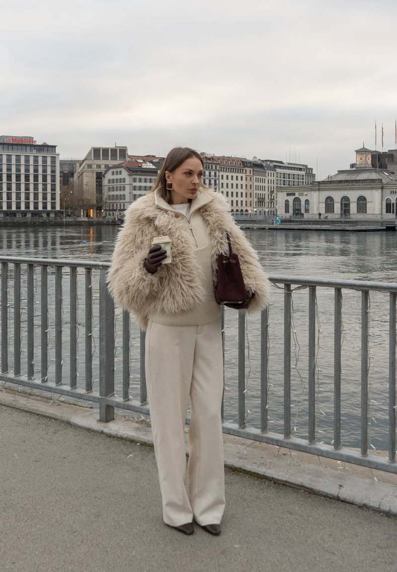 Veste en fausse fourrure beige, pull crème et pantalon large. Tenant une tasse de café, portant un sac à main bordeaux, debout près d'une rivière.