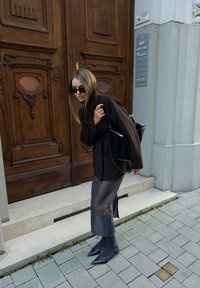 Brązowy, oversize'owy żakiet, przezroczysta szara spódnica, spiczaste czarne botki i czarna torba. Okulary przeciwsłoneczne dopełniają stylizację. Drewniane drzwi w tle.