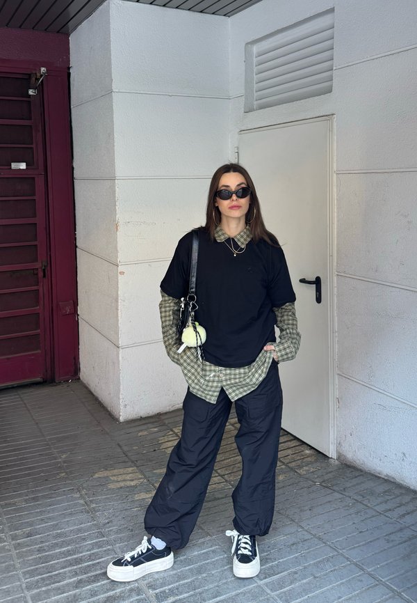 Ung kvinde iført solbriller, sort oversized t-shirt over grøn ternet skjorte, sorte bukser og sneakers, står i en flisebelagt korridor med hvide vægge.