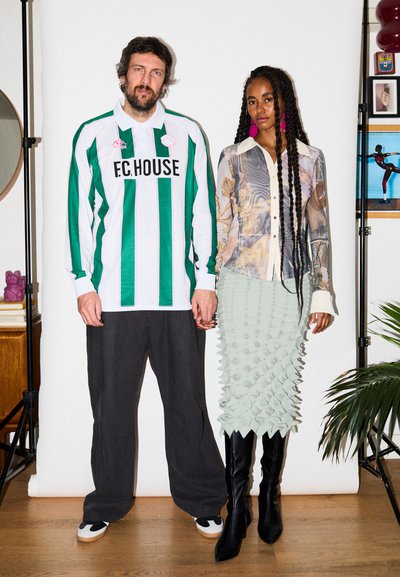 Hombre y mujer de pie, cogidos de la mano; el hombre lleva una camisa a rayas verdes y blancas y pantalones negros, la mujer lleva una blusa estampada y una falda texturizada con botas.