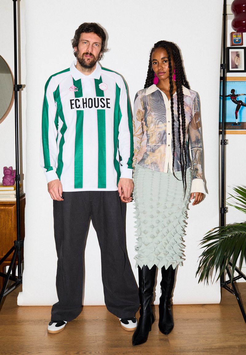 Άνδρας και γυναίκα στέκονται κρατώντας τα χέρια· ο άνδρας φοράει πράσινο και άσπρο ριγέ πουκάμισο και μαύρο παντελόνι, η γυναίκα φοράει μοτίβο μπλούζα και υφασμάτινη φούστα με μπότες.