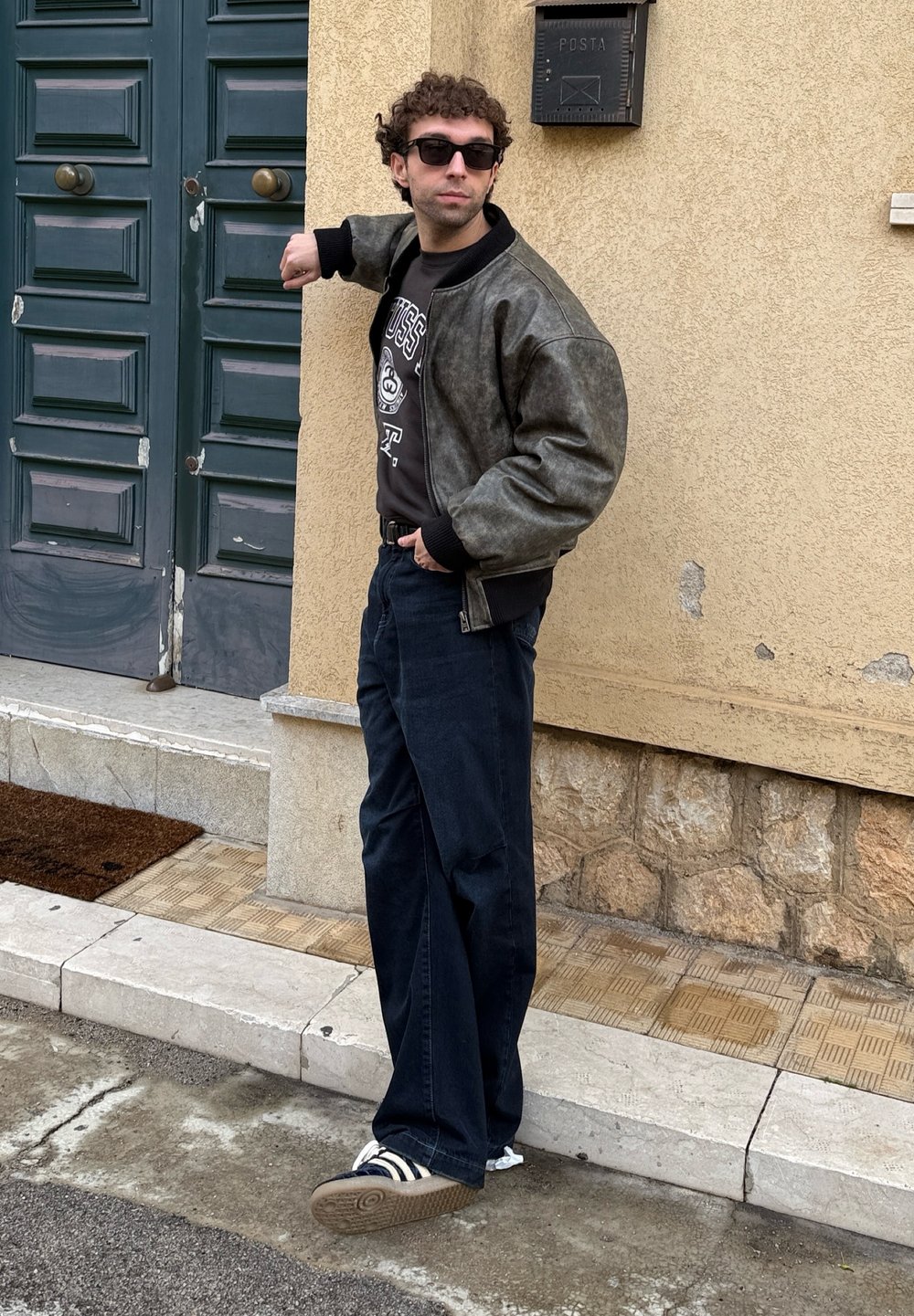 Άντρας με ηλιοθεραπευτικά γυαλιά και δερμάτινο μπουφάν στέκεται δίπλα σε μια μεγάλη πράσινη πόρτα, ακουμπώντας με το ένα χέρι στον τοίχο.