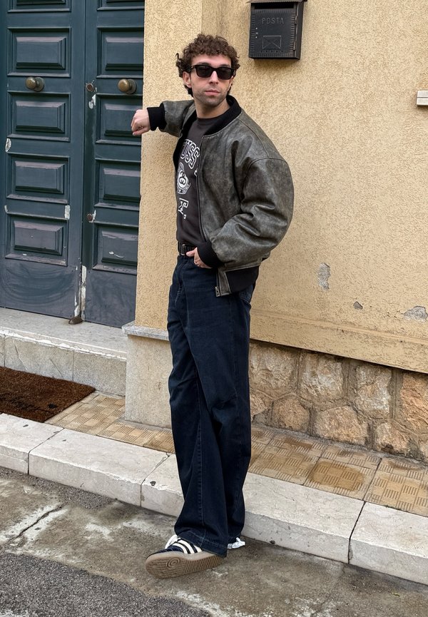 Uomo con occhiali da sole e giacca di pelle in piedi accanto a una grande porta verde, appoggiato con un braccio esteso al muro.