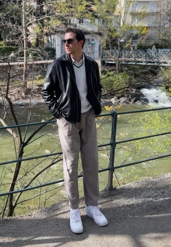 Schwarze Lederjacke über einem weißen Pullover mit grünem Streifen, graue Hose und weiße Sneaker. Steht neben einem Fluss.