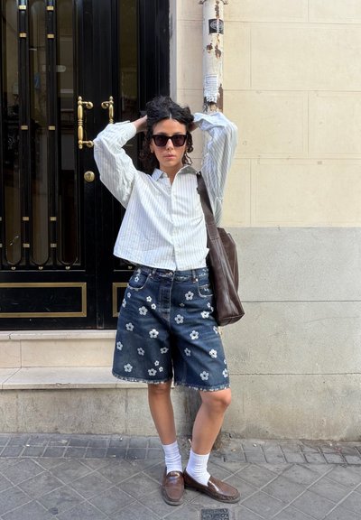 Camisa blanca y azul a rayas, pantalones cortos de mezclilla oscuros con bordados florales, bolso de piel marrón, calcetines blancos y gafas de sol oscuras frente a una puerta negra.