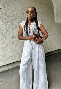 Δαντέλα μπλούζα με σχέδιο κλειδώματος, συνδυασμένη με χαλαρούς, ριγέ μπεζ παντελόνες. Τα αξεσουάρ περιλαμβάνουν μια καφέ τσάντα και γυαλιά ηλίου.