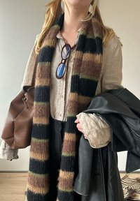 Gestreifter Schal in Schwarz-, Braun- und Grüntönen; weiche Textur. Dunkle Lederjacke und braune Tasche in den Händen gehalten. Hellbluse darunter.