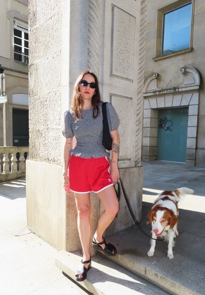 Pantalones cortos deportivos rojos con ribete blanco, una camiseta gris con un patrón, y sandalias de cuña negras. Un perro con correa está de pie junto a la persona, apoyado contra un pilar de piedra.