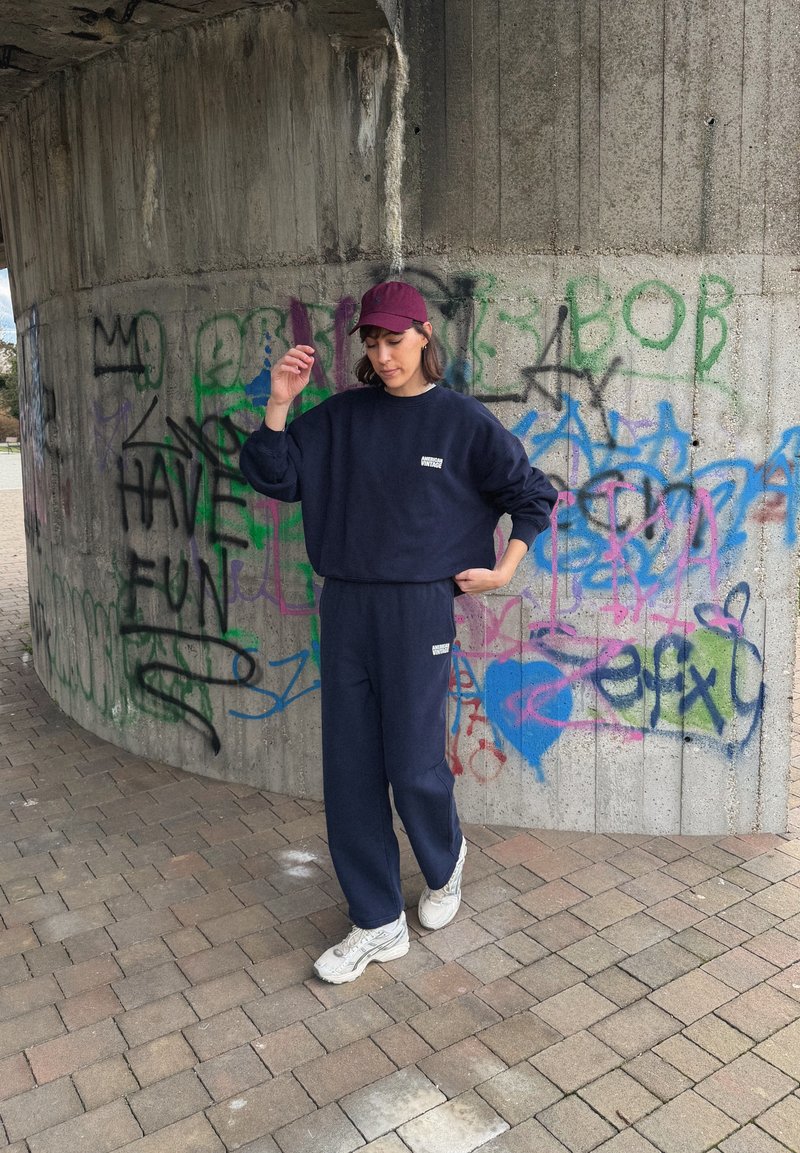 Donna con tuta da ginnastica blu navy e berretto color bordeaux che posa vicino a un muro di cemento coperto da graffiti colorati, inclusa la frase "DIVERTITI."
