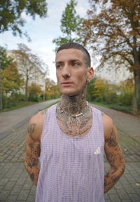 Jauns vīrietis ar tetovējumiem uz kakla un rokām, kurš valkā lavandas krāsas Adidas bezpaliktni un sudraba kaklarotu, stāv kokos apstādītā trotuārā.