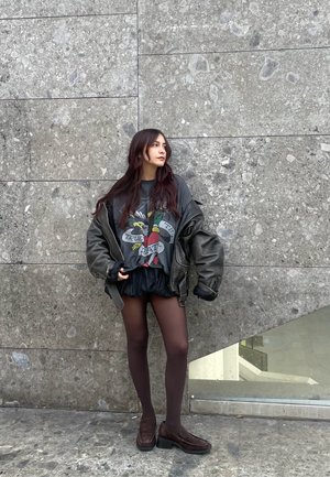 Chaqueta negra de cuero oversize, camiseta gráfica con diseño colorido, falda negra, medias transparentes y zapatos de slip oscuro. Fondo de piedra gris.