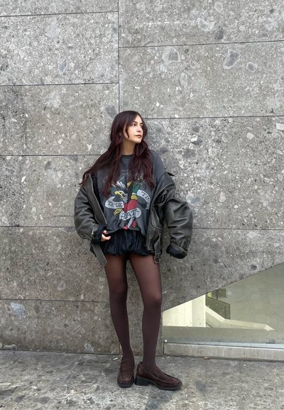 Chaqueta negra de cuero oversize, camiseta gráfica con diseño colorido, falda negra, medias transparentes y zapatos de slip oscuro. Fondo de piedra gris.