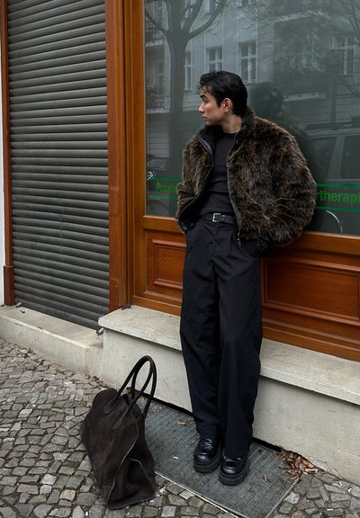 Chaqueta de piel sintética y cuero, suéter negro de cuello alto, pantalones anchos negros, zapatos negros robustos y un bolso de ante marrón sobre adoquines, cerca de una ventana de madera.