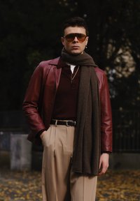 Bordeaux leren jack over een bruine gebreide sjaal, gecombineerd met een donkerrode polo, lichtbeige broek en een zwarte riem met zilveren hardware.