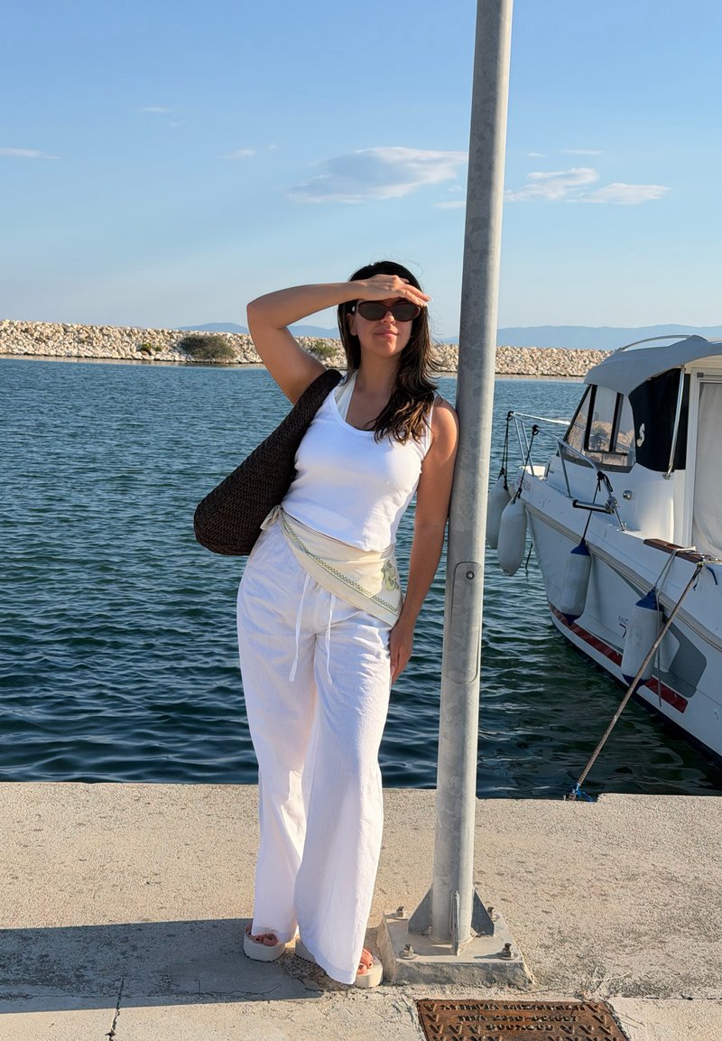 Γυναίκα με λευκή ενδυμασία και γυαλιά ηλίου που σκιάζει τα μάτια της, στέκεται σε προβλήτα κοντά σε βάρκα και ήρεμο νερό κάτω από καθαρό ουρανό.