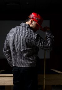 Homme portant une veste à motifs et une casquette rouge, tenant une queue de billard, debout à côté d'une table de billard en bois sous un éclairage tamisé.